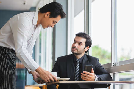 Asian barista man serving coffee for businessman in coffee shop. man sitting waiter coffee in cafe.の写真素材