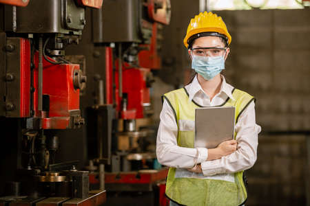 woman engineering wear protection face mask and safety helmet holding tablet standing front machine...