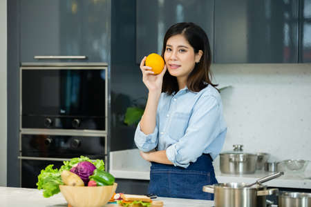 happy asian young woman holding orange, fruits and vegetables on table in kitchen at home.の写真素材