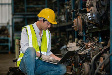 asian engineering man worker wear uniform and helmet using laptop checking automotive parts in warehouse. workplace factory industrial heavy machine.の写真素材