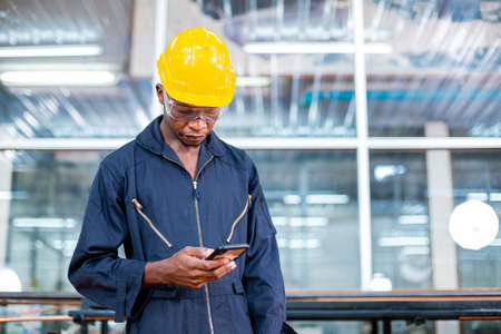 Engineering male african american workers wear soundproof headphones and yellow helmet using work smartphone. factory industrial concept.の写真素材