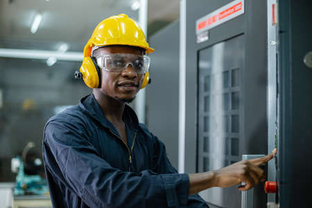 Engineering male african american workers wear soundproof headphones and yellow helmet working at operating CNC machine. work factory industrial concept.の写真素材