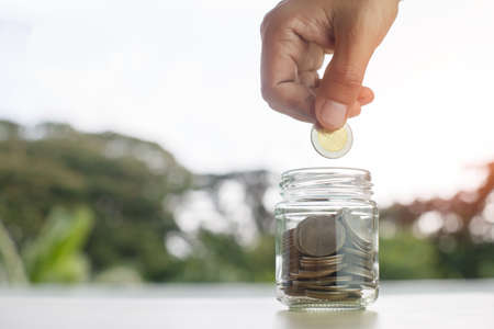 Coins in glass bottle put on table with hand putting money coins nature background, growth save or...
