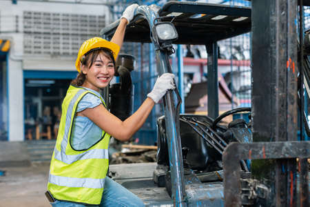 women wear safety helmet happy smiling looking at camera driver forklift warehouse in factory. Female worker industrial operate. control loading container box. logistics export import concept.の写真素材
