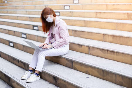 Asian young woman wearing face mask protection and sitting on floor and working using on laptop. Work outdoor.の写真素材