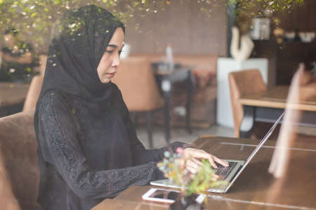 Asian muslim in black headscarf woman working with laptop in cafe morning time.の写真素材