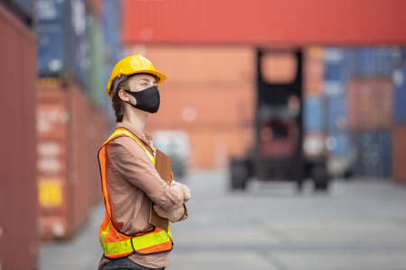 women foreman wear protection face mask stand hand cross with clipboard at loading containers in shipping yard, for logistics and import export company. business logistics concept.の写真素材