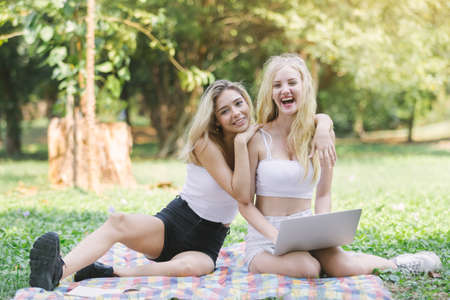 Two woman blonde hair friends embrace sitting smile and laugh with laptop in green park, Woman friendship.の写真素材