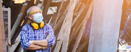 Portrait asian senior man carpenter crossed hands wear protective mask dust and headphone standing at woodwork.の写真素材