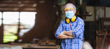 Portrait asian senior man carpenter crossed hands wear protective mask dust and headphone standing at woodwork.の写真素材