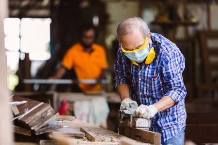 asian senior man carpenter wear protective mask dust and headphone working with electric planer In...