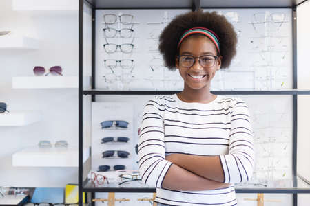 Portrait happy young woman African American afro hair smile wear spectacles crossed arm standing at in optical shop. modern ophthalmologist concept.の写真素材