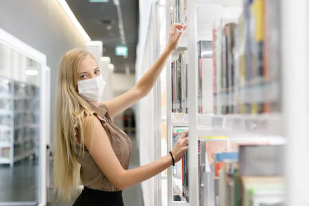 Beautiful woman with blond hair wear medical mask standing bookshelf at library, Education concept.
