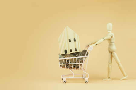 Wooden mannequin or puppet pushing shopping cart with wooden house and coins money on orange  background, business sale and purchase property concept.の写真素材