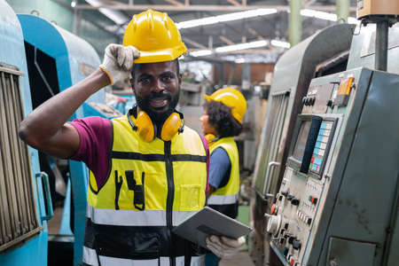 Engineering male african american happy smiling workers wear soundproof headphones and yellow helmet holding tablet working at operating CNC machine. work factory industrial concept.の写真素材
