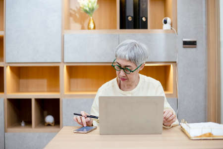 asian elderly female gray hair wear spectacles working on laptop computer at home office. working...