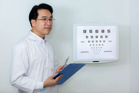 Man ophthalmologist wear uniform holding clipboard standing near eye chart vision test in clinic.
