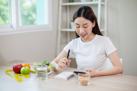 asian females using smartphones and taking notes eating healthy food each day with vegetables fruit on table. health and care concept.の写真素材