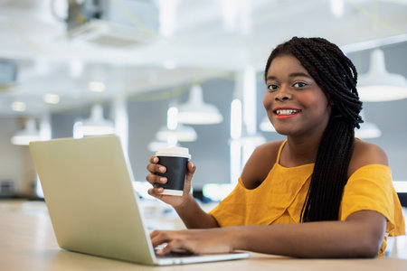 Happy young businesswoman African American cheerful smiling looking at camera use laptop holding coffee cup. female working successful.の写真素材