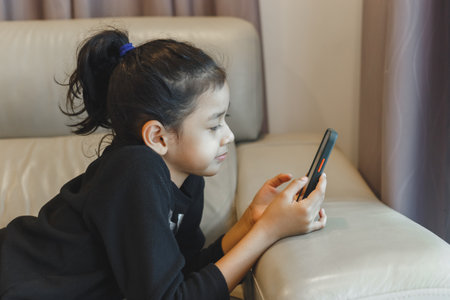 Child girl lie down and play looking smartphone study online. little girl chatting online learning. Little girl on screens, social media addiction.の写真素材