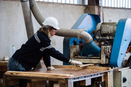 Woman wearing safety uniform and hard hat working on wood sanding electric machines at workshop...