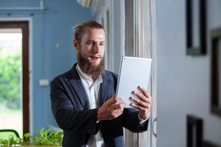 Portrait handsome businessman with beard holding tablet standing at side window office. happy smart man mature wear suit.の写真素材
