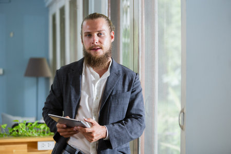 Portrait handsome businessman with beard holding tablet standing at side window office. happy smart man mature wear suit.の写真素材