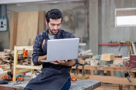 young carpenter caucasian man with laptop make drawing on wood to design at workspace. craftsman profession in wood factoryの写真素材