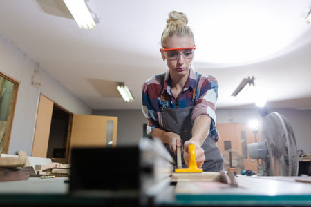 carpenter woman wear uniform and goggles working use electronic saw cutting wood. craftsman profession in wood factory. woodworking industry.の写真素材