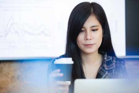 Asian businesswoman holding coffee cup using laptop computer, working at office. Female office...