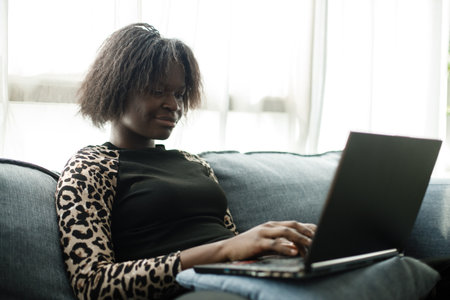 young woman african american using laptop on sofa in modern house living room. Using computer to talk and buy things shop online. playing watching movie via internet.の写真素材