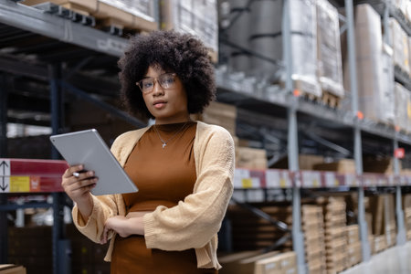 Young African American female looking at camera working in warehouse using digital tablet checking inspection on shelves.woman worker check stock inspecting in storage logistic factory.の写真素材