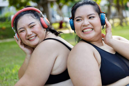 Cheerful laugh smiling friends young asian woman happy wear headphone listen to music in sportswear walking after exercise. Two sporty women body curvy positive having fun workout in garden.の写真素材