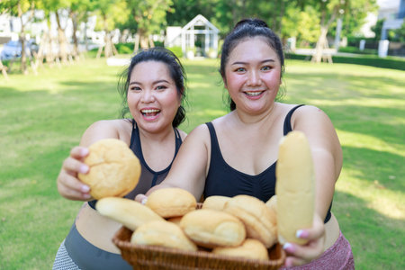 Cheerful two woman in sportswear have fun and enjoy eating bread while exercising in garden. Asian girl holding bread looking at camera standing in the park.の写真素材