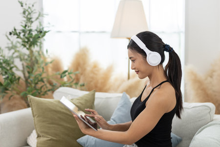 Young women wearing headphones is engrossed in a tablet, seated comfortably in her living room. The space is bright and airy, adorned with minimal decor and soft furnishings.の写真素材