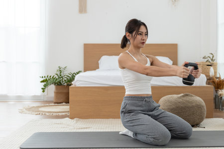 Young women engages in strength training using a fitness device while kneeling on a mat in a cozy and well-lit bedroom. The environment promotes wellness and exercise.の写真素材