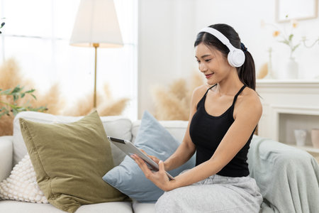 Young women wearing headphones is engrossed in a tablet, seated comfortably in her living room. The space is bright and airy, adorned with minimal decor and soft furnishings.の写真素材