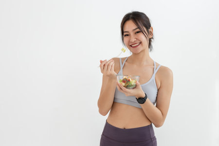 Asian attractive lady sportswear healthy eating salad vegetables over white background. Female holding salad bowl various vegetables and fruit healthy food for weight loss and exercise.の写真素材