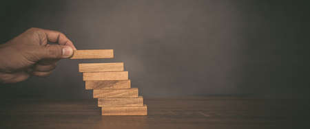Close-up hand is placing wood block tower stacked in stair step with caution to prevent collapse or...