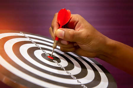 Hand holding red dart arrow  hitting in the target center of dartboard on bullseye.の写真素材