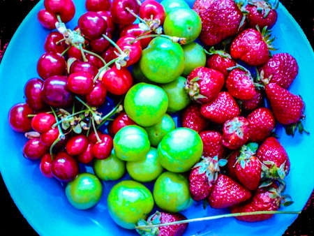 Red and green cherries and strawberries in a blue bowl on a black backgroundの写真素材