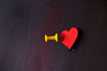 A red heart is pinned with a yellow button to a multi-colored surface covered with water dropsの写真素材