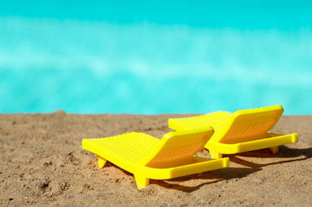 Two yellow toy sunbeds stand on the sand on the beach against the backdrop of the blue water of the ocean or sea on a hot sunny dayの写真素材