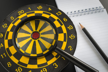 aiming target analysis concept with magnifying glass on dart board, pencil and note paper.