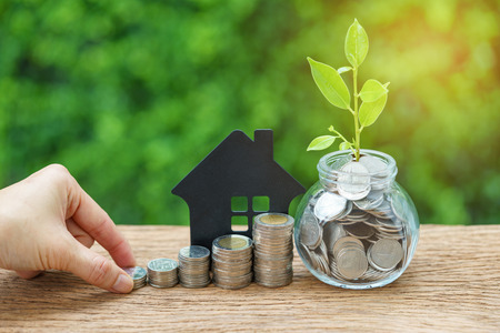 growth sprout plant in jar with full of coins and hand holding stack of coins with paper house as...