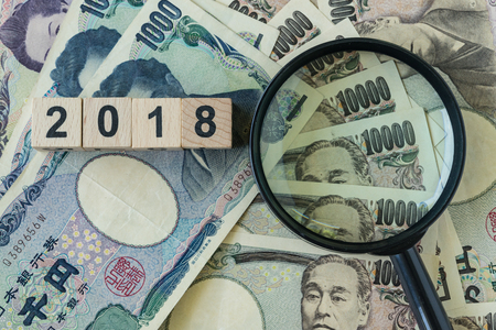 Wooden cube with number 2018 and magnifying glass on pile of japanese yen banknotes as financial safe haven or goals and target concept.の写真素材