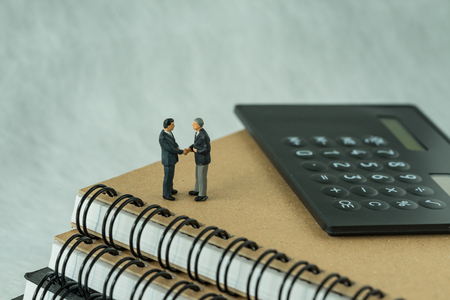 Miniature people: Small figure businessmen handshaking and standing on notebook with calculator as business agreement concept.の写真素材