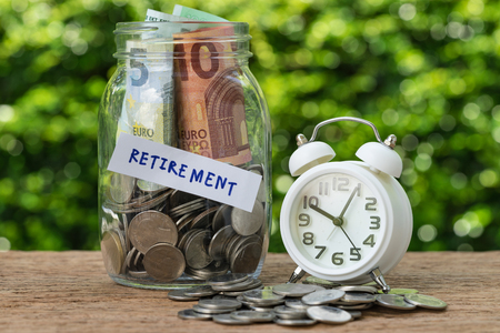 glass jar bottle labeled as retirement with full of coins and banknotes, white alarm clock as savings or investing for retirement concept.の写真素材