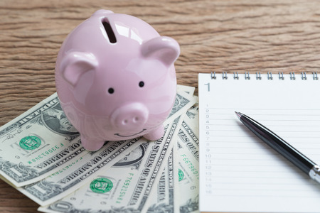 Financial goals, expense or saving write down, pen on paper notepad with number listed and smile pink piggy bank on money US dollar banknotes on wooden table.の写真素材
