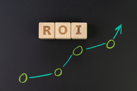 Return on Investment, ROI, performance measure of business effeiciency, target and goal, cube wooden block with word ROI with pastel green hand writing graph on chalkboard.の写真素材